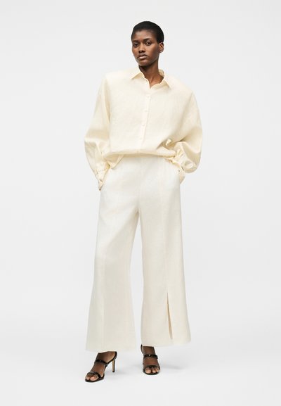 Γυναίκα φορώντας μια φαρδιά κρεμ πουκάμισο με κουμπιά και ταιριαστό παντελόνι φαρδύ με σχισμές, συνδυασμένο με μαύρα πέδιλα με τακούνι, στέκεται με τα χέρια στις τσέπες.