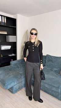 Schwarzes Langarmoberteil mit goldenen Knöpfen, kombiniert mit weit geschnittenen grauen Hosen. Accessoires sind schwarze Sonnenbrillen und eine kleine Handtasche.