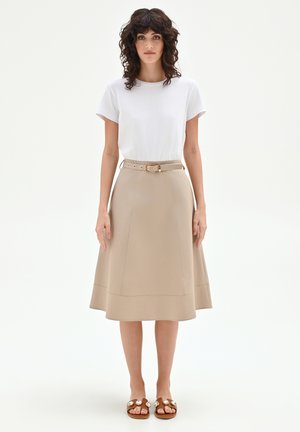 Femme aux cheveux bouclés foncés portant un haut blanc à manches courtes, une jupe trapèze beige ceinturée et des sandales marron ornées de bijoux décoratifs, debout.