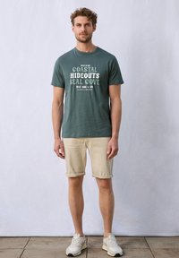 Grön bomullströja med vit grafisk text, kombinerad med beige upprullade shorts. Modellen bär ljusa sneakers och står mot en neutral bakgrund.