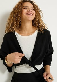 Cárdigan negro abierto en la parte delantera con tejido texturizado, usado sobre una camiseta blanca de canalé con un nudo en la parte delantera. Brazaletes dorados en la muñeca.