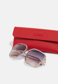 Lunettes de soleil avec une monture transparente, des verres dégradés du violet clair au foncé, accompagnées d'un étui en cuir rouge texturé embossé du logo "GUESS".