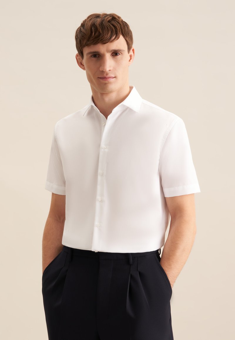 Homme portant une chemise blanche à manches courtes boutonnée avec un pantalon noir, debout les mains dans les poches devant un fond beige uni.