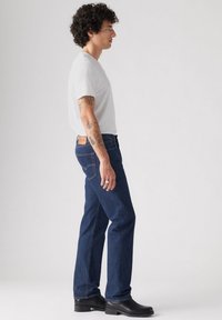 Jeans azul oscuro de corte recto, acompañados de una camiseta blanca ajustada. Presenta un parche de cuero en la parte posterior y botas negras hasta el tobillo.