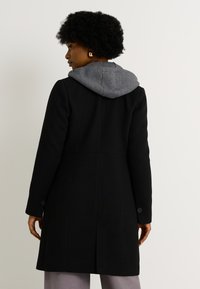 Abrigo de mezcla de lana negro con capucha gris, diseño ajustado, puños con botones y costura en la parte posterior. Textura suave, silueta moderna.