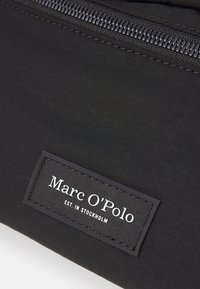 Juodas audinio maišelis, turintis lygią tekstūrą ir matomą užtrauktuką. Stačiakampė etiketė baltais rašmenimis skelbia „Marc O'Polo EST. IN STOCKHOLM“.