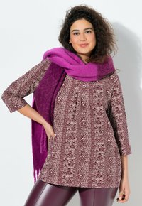 Langärmlige burgunderfarbene Tunika mit weißem Blumenmuster, kombiniert mit einem dicken lila Schal und glänzenden burgunderfarbenen Leggings. Strukturierter Stoff.