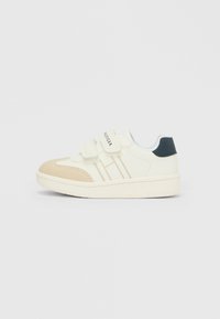 Scarpetta da bambino bianca e beige con due cinturini in velcro e toppa sul tallone blu navy su uno sfondo chiaro uniforme.