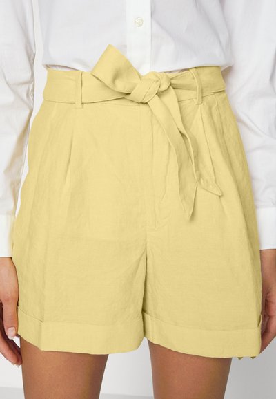 Calções amarelos de linho com um cinto amarrado na cintura, pernas largas e bainhas dobradas. Combinados com uma camisa branca de botões. A textura é ligeiramente amarrotada.