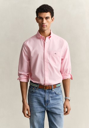Joven con camisa rosa claro abotonada con las mangas remangadas, vaqueros azules, cinturón marrón y reloj de pulsera plateado, de pie frente a un fondo liso.