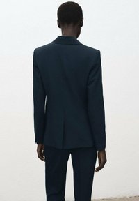 Donker marineblauw pak met een getailleerde pasvorm, voorzien van een notch-revers, een enkele achterkant met een split en een gestructureerde stof. Details bevatten knopen aan de manchetten.