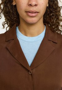 Giacca marrone con bottoni in tessuto strutturato, con collo a scalpello. Indossata sopra una maglietta a maniche corte azzurro chiaro.