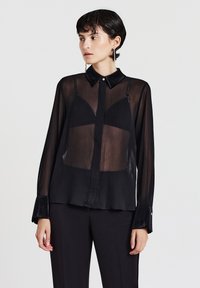 Camicia nera trasparente con scollo con colletto, chiusura con bottoni sul davanti e maniche lunghe con polsini in velluto. Tessuto strutturato con leggera trasparenza.
