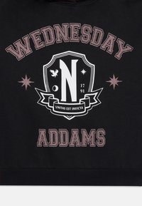 Hoodie noir portant "WEDNESDAY" et "ADDAMS" en texte rose. Le graphique central comprend un bouclier avec un "N", des contours blancs et des accents d'étoiles.