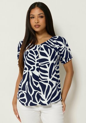 Mujer con cabello largo y oscuro que lleva una blusa abotonada de manga corta con patrón abstracto en azul marino y blanco, y pantalones blancos.