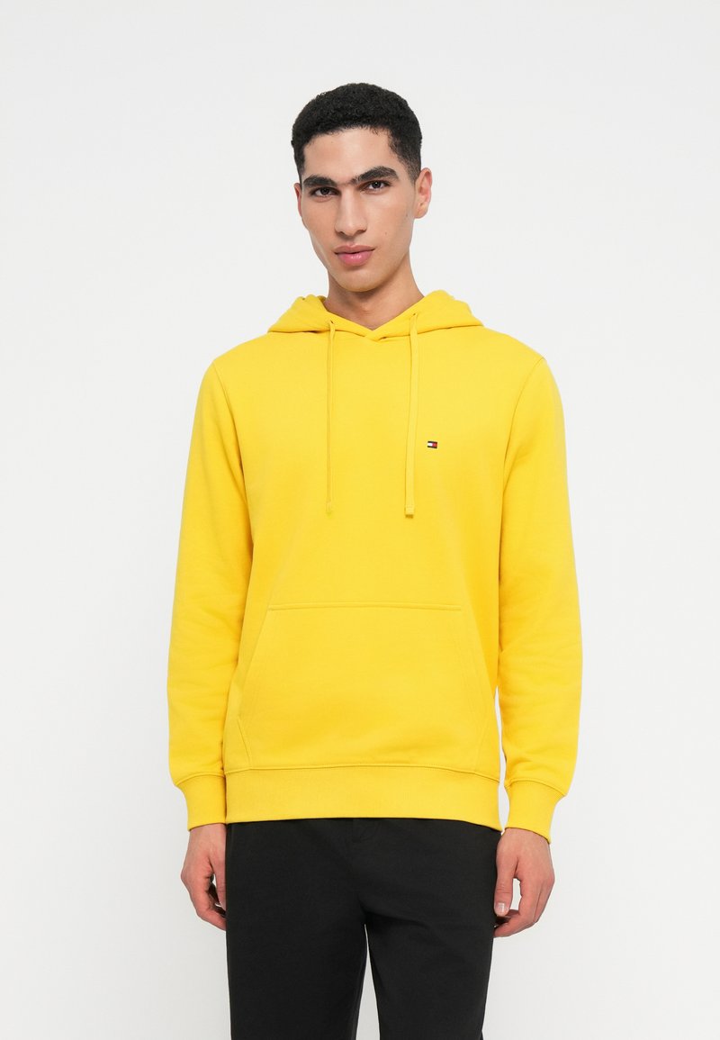 Mladý muž oblečený v žiarivo žltom mikinovom svetri s prednou kapsou a šnúrkami, v kombinácii s čiernymi nohavicami, stojaci na bielom pozadí.