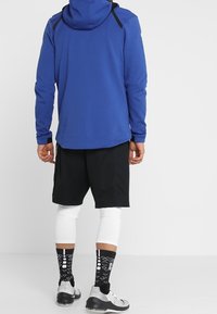 Sudadera azul con capucha y detalles en negro, combinada con pantalones cortos negros, mangas de compresión blancas y calcetines negros con estampado. Nota: calzado deportivo.