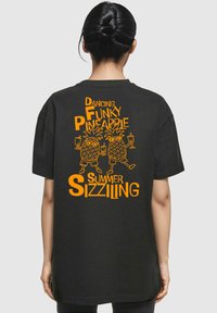 Sorte bomulds t-shirt med et orange grafisk motiv af to dansende ananasser med teksten "Dancing Funky Pineapple Summer Sizziling" ovenover og nedenfor.