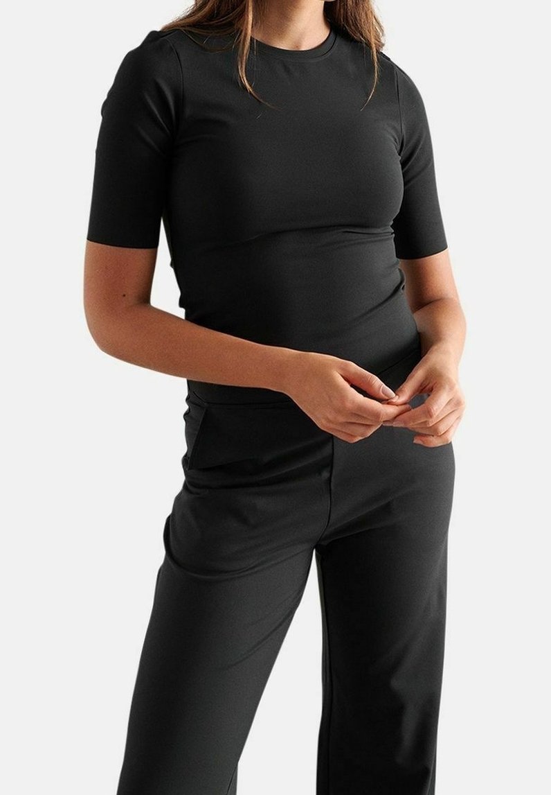 Femme portant un haut noir ajusté à manches courtes et un pantalon noir, debout les doigts entrelacés à la taille devant un fond uni.