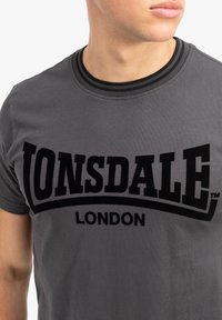 Pelēks kokvilnas T-krekls ar izteiktu melnu "Lonsdale London" logotipu. Ribota melna apaļā kakla izgriezums. Gluda tekstūra, ikdienišķs dizains.