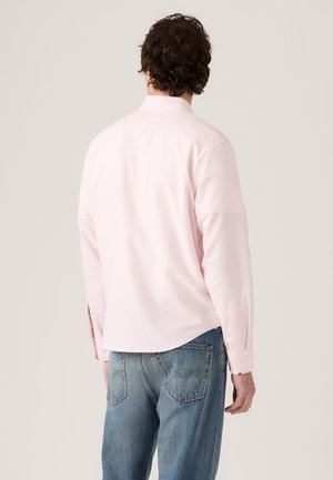 Homme aux cheveux bouclés et foncés portant une chemise rose clair à manches longues et un jean bleu, vu de dos sur un fond uni.