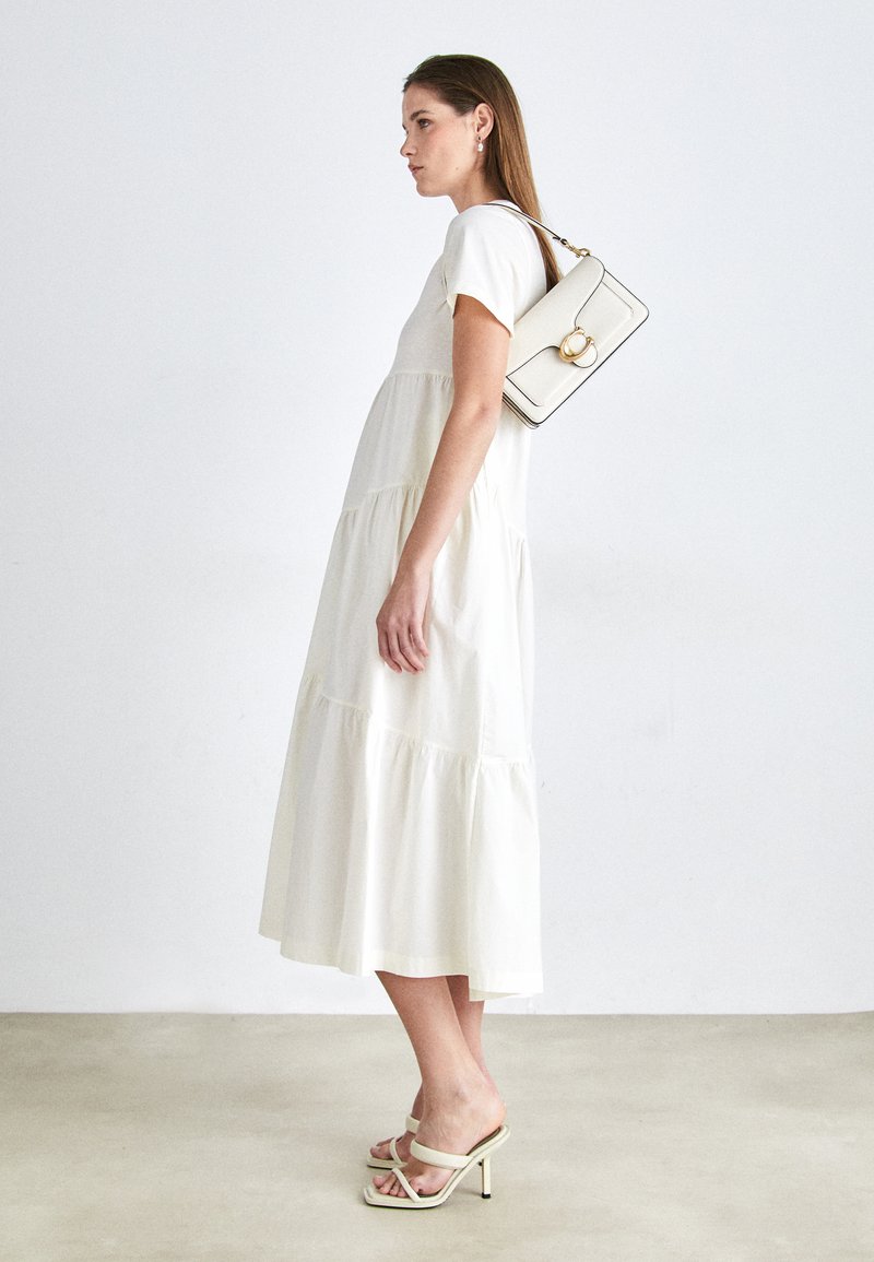 Biała, warstwowa sukienka o luźnym kroju z krótkimi rękawami. Modelka niesie kremową torebkę z ozdobnym złotym pierścieniem i nosi jasne sandały na obcasie z paskami.