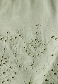 Un tessuto di colore verde chiaro presenta un ricamo intricato con aperture circolari e motivi floreali, evidenziando cuciture delicate e una superficie liscia.