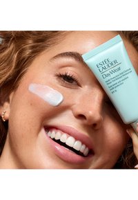 Hellblauer Estée Lauder DayWear-Tube, der neben dem Gesicht eines Modells gehalten wird, mit einem weißen Cremefleck auf ihrer Wange und einer glatten, strahlenden Hautstruktur.