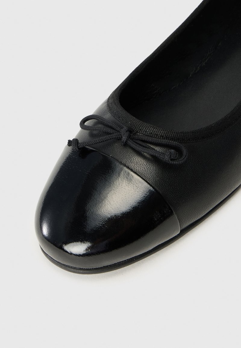 Ballet flat noire avec un bout arrondi, présentant un cap en cuir verni noir brillant et un dessus en cuir mat, agrémenté d'un nœud noir.