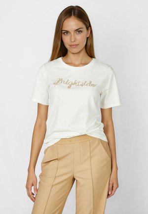 Woman wearing white t-shirt with "Brightside" text in gold beads paired with high-waisted beige trousers, standing against plain background.