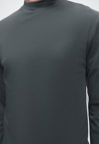 Gros plan d'une personne portant un t-shirt à col roulé à manches longues gris foncé uni sur un fond neutre.