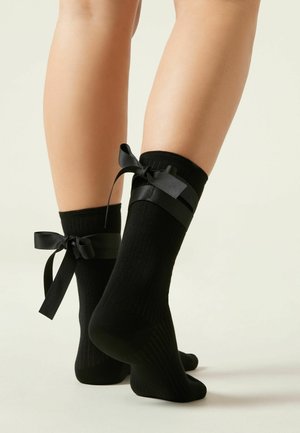 Schwarze gerippte Socken mit satinähnlichen Bändern, die oben gebunden sind, und einem weichen Material, das einen engen Sitz am Knöchel bietet.