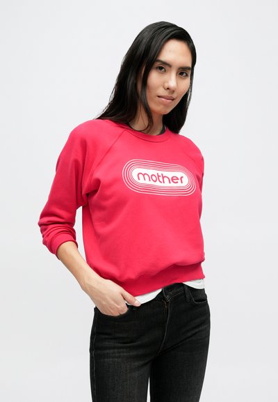 Raudonas džemperis su balta grafine „mother“ logotipo spauda, apvalia iškirpte ir ribiniais rankogaliais, derinamas su tamsiais džinsais iš džinso audinio.