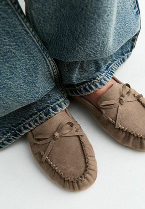 Bruine suède mocassinslippers met strikdetails gedragen met blauwe spijkerbroek op een lichte ondergrond.