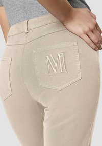 Beige jeans gemaakt van een gladde, rekbare stof met achterzakken. De rechterzak heeft een subtiele geborduurde "M" logo.
