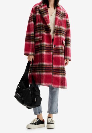 Personne portant un long manteau à carreaux rouge et noir, jean bleu court, baskets noires et blanches, tenant un sac noir avec plusieurs poches.