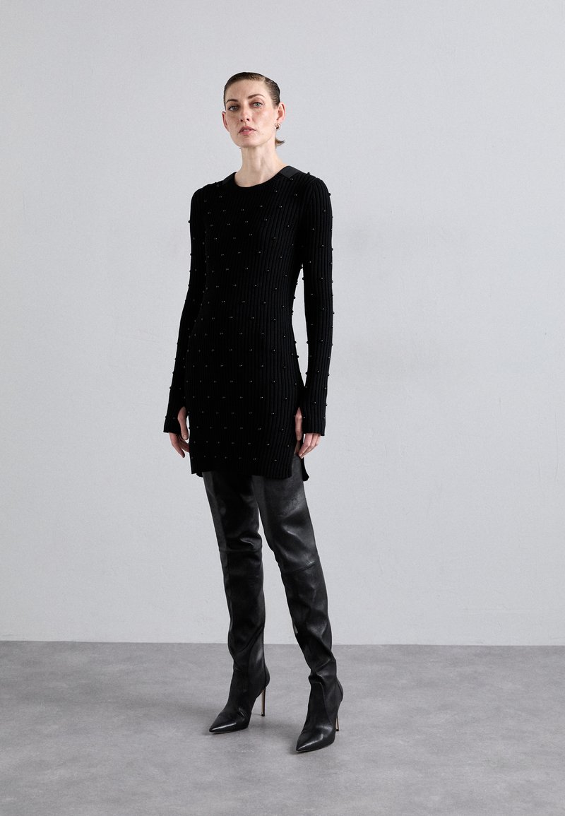 Robe noire côtelée avec des détails texturés et de petites accents, associée à des cuissardes en cuir noir ajustées et à des talons aiguilles.