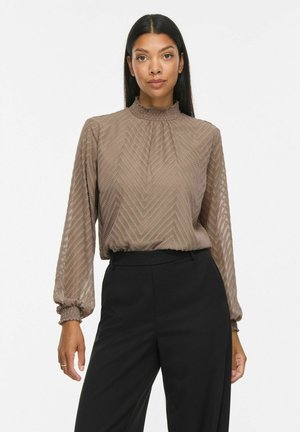 Femme aux longs cheveux noirs portant un chemisier marron texturé à manches longues et un pantalon noir taille haute, debout sur fond blanc.