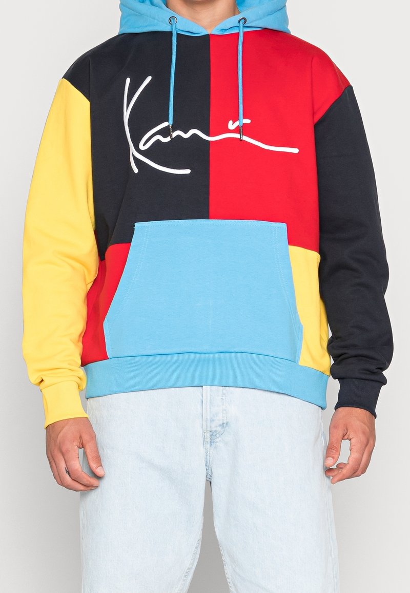 Sudadera con capucha colorida hecha de una mezcla de algodón. Presenta paneles en azul, rojo, negro y amarillo, con un bolsillo estilo canguro y un detalle de firma en blanco.