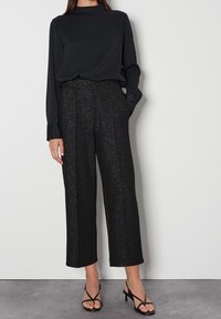 Kvinna iklädd en svart långärmad topp, högmidjade glittriga svarta capribyxor och svarta snöriga klackar, stående mot en enfärgad vägg.