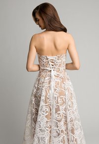 Suknia bez ramiączek z haftowanymi wzorami kwiatowymi w białym kolorze na przezroczystym, beżowym tle. Zapięcie na plecach z koronkowymi detalami. Długa, zwiewna spódnica.