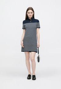 Marineblau-weiß gestreiftes Polo-Kleid aus Baumwolle, mit einem Kragen und drei goldenen Knöpfen sowie kurzen Ärmeln. Kombiniert mit schwarzen Slippern.