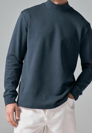 Mand iført en mørkeblå rullekrave og lyse beige bukser, der står med den ene hånd i lommen mod en ensfarvet grå baggrund.