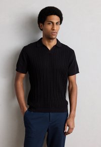 Μαύρο πουλόβερ πόλο με κοντά μανίκια, με υφασμένο σχέδιο πλέξης καλωδίου και γιακά σε σχήμα V. Σε συνδυασμό με μπλε παντελόνια.