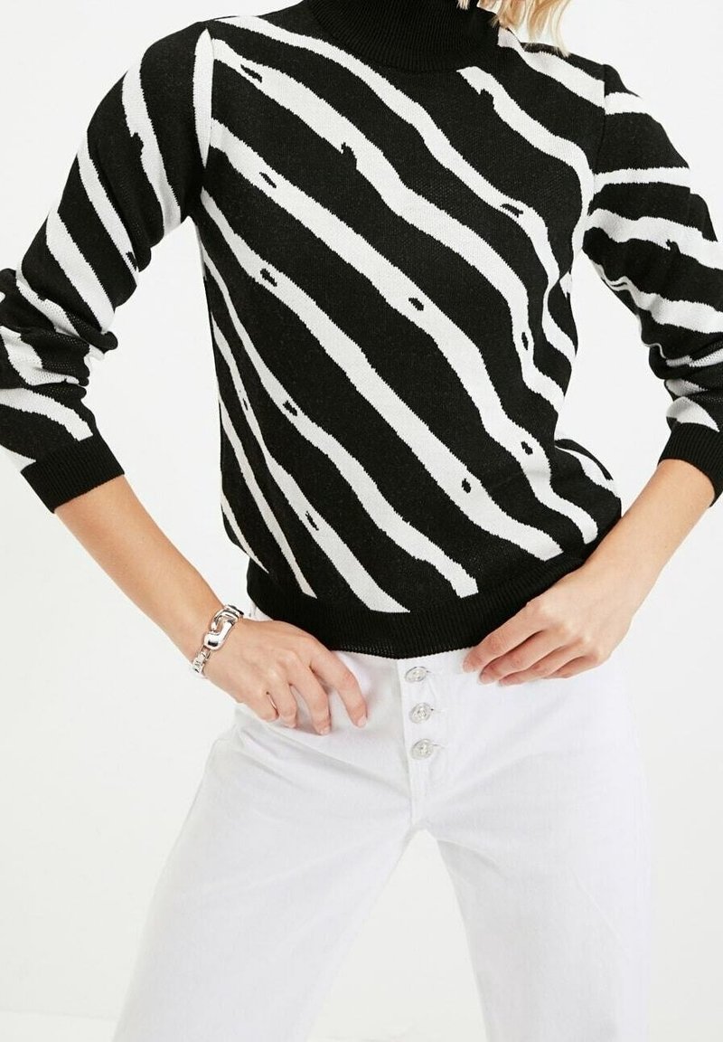 Musta-valkoraidallinen neulepaita korkealla kaula-aukolla ja resorireunuksilla, puettu valkoisten korkeavyötäröisten farkkujen päälle, joissa on nappikoristeet.