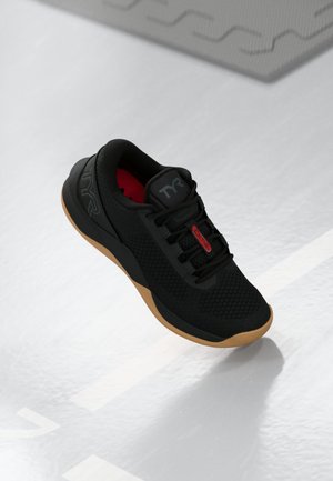 Black athletic shoe with gum sole, red "CXT2" tag on laces, and "TYR" logo, placed on a light gray sports court floor.