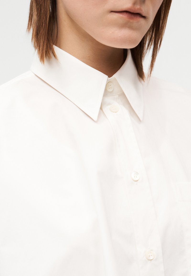 Chemise blanche à boutons avec un col structuré, présentant une texture de tissu lisse et une poche poitrine unique. Quatre boutons visibles.