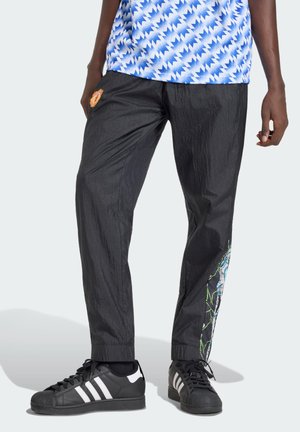 Osoba koja nosi crne Adidas tenisice, crne naborane hlače s šarenim uzorkom sa strane i plavu košulju s geometrijskim uzorkom.