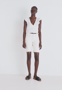 Un top blanc court avec des manches volantes, associé à un short en denim crème pâle avec un ourlet effiloché. Des sandales noires avec des accents cloutés.