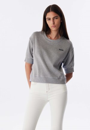 Pull en coton gris à manches courtes avec des poignets et un ourlet côtelés, arborant un petit logo brodé sur la poitrine gauche. Porté avec un jean blanc.